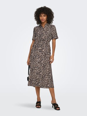 Vrouw die een midi-jurk met luipaardprint en knopen draagt, een kleine zwarte handtas vasthoudt en zwarte sandalen met hakken, staand tegen een effen achtergrond.