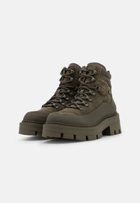 Botas de senderismo de ante en verde oliva oscuro con suelas de tacos gruesos y ganchos metálicos en los cordones, mostradas una al lado de la otra sobre un fondo liso.