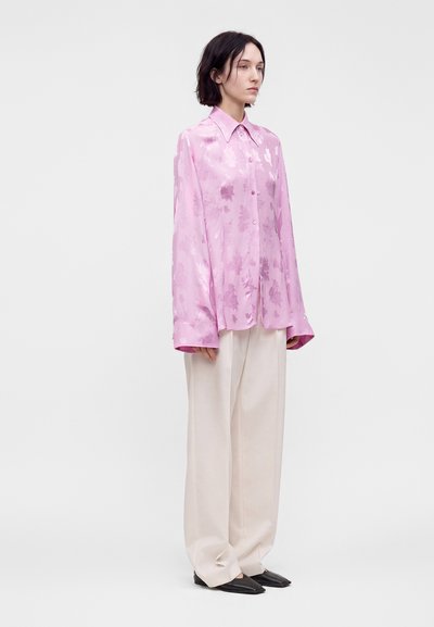 Femme portant une chemise en satin fleurie rose pâle oversized, un pantalon large beige et des chaussures noires pointues, debout sur un fond clair uni.