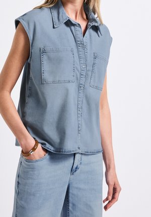 Sleeveless light blue denim shirt with front pockets and snap buttons worn with matching light blue jeans and wooden bracelet on left wrist.