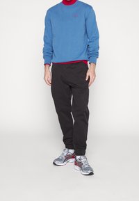 Sudadera azul con cuello redondo, acento rojo debajo, pantalones negros y zapatillas deportivas grises con detalles rojos y blancos.