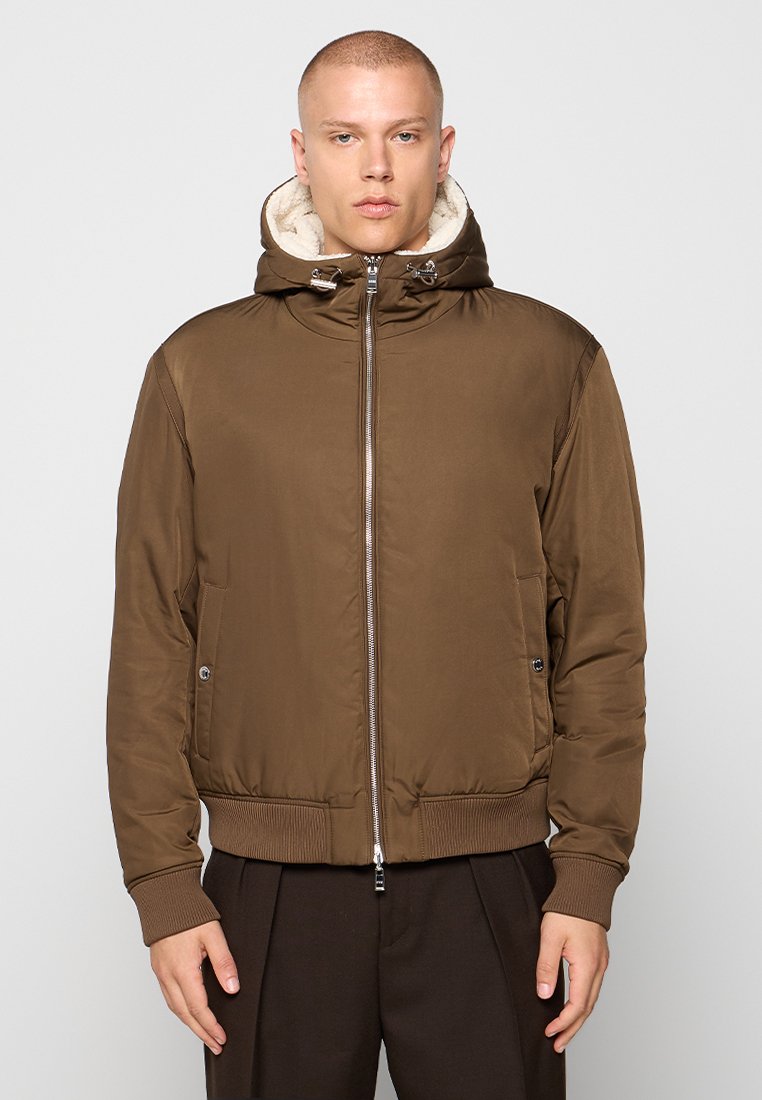 Veste matelassée marron avec une fermeture éclair, une capuche et une doublure en polaire beige contrastante. Dotée de poches latérales et de poignets côtelés.