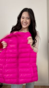 Lächelnde Frau mit langen dunklen Haaren hält eine leuchtend pinke, gesteppte Jacke vor einer schlichten hellen Wand im Innenraum.