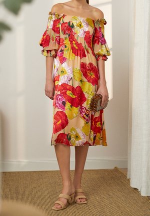 Vrouw draagt een off-shoulder jurk tot op de knie met rood, geel en roze bloemenpatroon, houdt een beaded clutch vast en draagt beige sandalen.