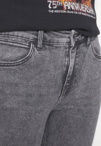 Los pantalones de mezclilla gris cuentan con un diseño clásico de cinco bolsillos, un sutil desgaste, una tela texturizada y costuras reforzadas en las uniones.