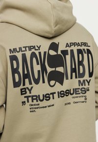 Sudadera beige con texto grande en negro en la parte de atrás: "MULTIPLY BACKSTAB’D BY MY TRUST ISSUES" con detalles de marca adicionales debajo.