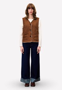 Frau in braunem Knopf-Westen über weißem Langarmshirt, mit weit geschnittenen, dunkelblauen Jeans mit großen Säumen und braunen Schuhen, stehend vor weißem Hintergrund.
