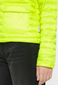 Lime green puffer jacket with a quilted design, featuring a chest pocket and a branded label on the sleeve, shown with black pants.