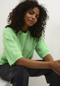 Lichtgroene gebreide trui met korte mouwen, voorzien van een bladpatroon; gedragen met donkergrijze broek, huidkleur zichtbaar op de armen.