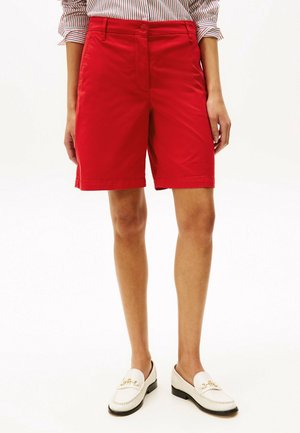 Donna con pantaloncini rossi vivaci fino al ginocchio, camicia a righe con maniche arrotolate e mocassini bianchi con dettagli dorati.