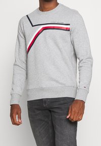 Mand iført en lysegrå sweatshirt med marineblå, hvid og røde skrå striber samt Tommy Hilfiger-logo, parret med mørkegrå jeans.