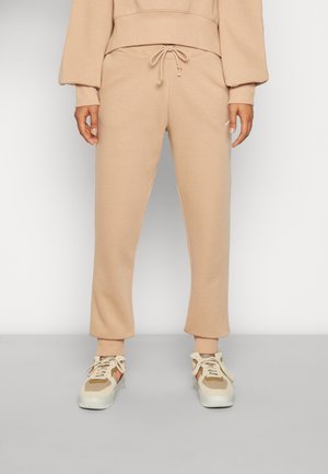 Persoon draagt beige sweatpants met trekkoord, omgeslagen enkels, en beige sneakers met witte zolen, staand tegen een effen achtergrond.