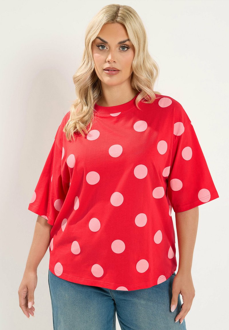 Femme blonde portant une chemise rouge ample avec de gros pois roses et un jean bleu, debout devant un fond clair uni.