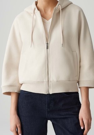 Personne portant un sweat à capuche crème zippé avec poignets et ourlet côtelés, associé à un jean bleu foncé, sur un fond neutre.
