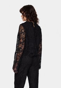 Top in pizzo nero con maniche lunghe, motivo floreale e scollatura alta. Presenta un'apertura a chiave sul retro e orlo a scallop, abbinato a pantaloni neri coordinati.