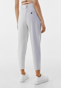 Světle šedé kalhoty s kombinací vertikálních proužků a strukturované, jednotné látky. Zadní kapsa s černým knoflíkovým detailem.