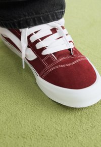Punainen mokkanahka sneaker valkoisilla nauhoilla ja sivusuorilla, matalalla varrella ja kumipohjalla, asetettuna vihreälle tekstuuripinnalle.