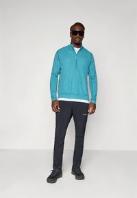 Maglione teal con cerniera, realizzato in tessuto leggero, abbinato a pantaloni neri con logo e tasche. Il modello indossa occhiali da sole e scarpe nere.