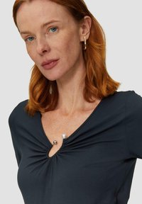 Marineblauwe top met een ronde halslijn, voorzien van plooien en decoratieve parel- en metaalaccenten in het midden. Zachte, gladde stof met korte mouwen.