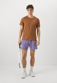 Camisola atlética de mangas curtas castanha com logótipo, combinada com calções lilás claros. O modelo segura uma raquete de ténis, vestindo sapatilhas brancas e meias.