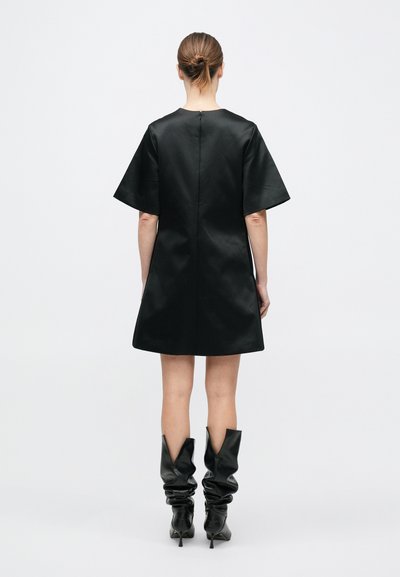 Robe en satin noir avec des manches courtes et larges et une coupe droite, dotée d'une fermeture éclair cachée au dos et d'une texture lisse et brillante. Portée avec de longues bottes noires froissées.