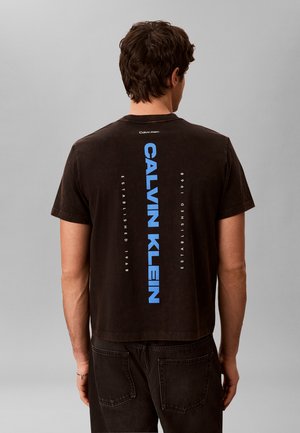 Homme aux cheveux courts et bruns portant un T-shirt noir Calvin Klein avec un grand logo vertical bleu et un jean noir, debout devant un fond gris.
