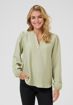 Blouse à manches longues, vert clair, avec un col en V, des poignets froncés et une texture douce. Associée avec un pantalon noir pour le contraste.