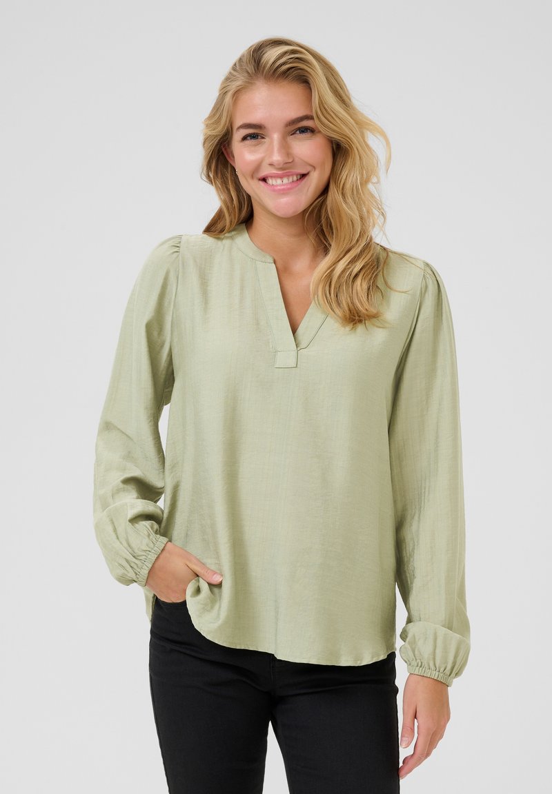Long-sleeve, light green blouse with a V-neck, gathered cuffs, and soft texture. Paired with black pants for contrast.
