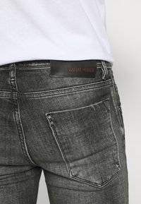Jeans de mezclilla negra deslavada con corte ajustado, que presentan un parche de cuero con la etiqueta "ANTHONY MORATO" y herrajes plateados en los bolsillos delanteros.