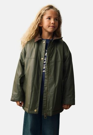 Grøn vandtæt jakke med brun krave, trykknapper og lynlås. Båret over en blå skjorte, parret med blå jeans.