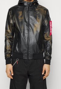 Man med en svart och brun spräcklig zip-up jacka med huva och en röd "Remove Before Flight"-tagg på vänster ärm.