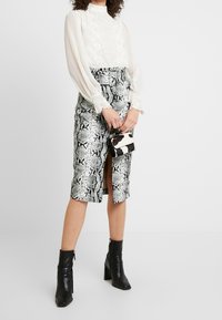 Kvinna klädd i en vit långärmad blus och en högmidjad blyertskjol med ormtryck, tillsammans med svarta ankelstövlar, håller en svart och vit väska.