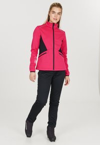 Fuchsia softshell-jakke med sorte accenter, høj krave, frontlynlås og sidelommer. Kombinationen med sorte tætsiddende bukser og sort fodtøj.