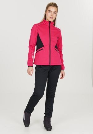Fuchsia softshell jas met zwarte accenten, hoge kraag, voorrits en zijzakken. Gecombineerd met zwarte strakke broek en zwarte schoenen.