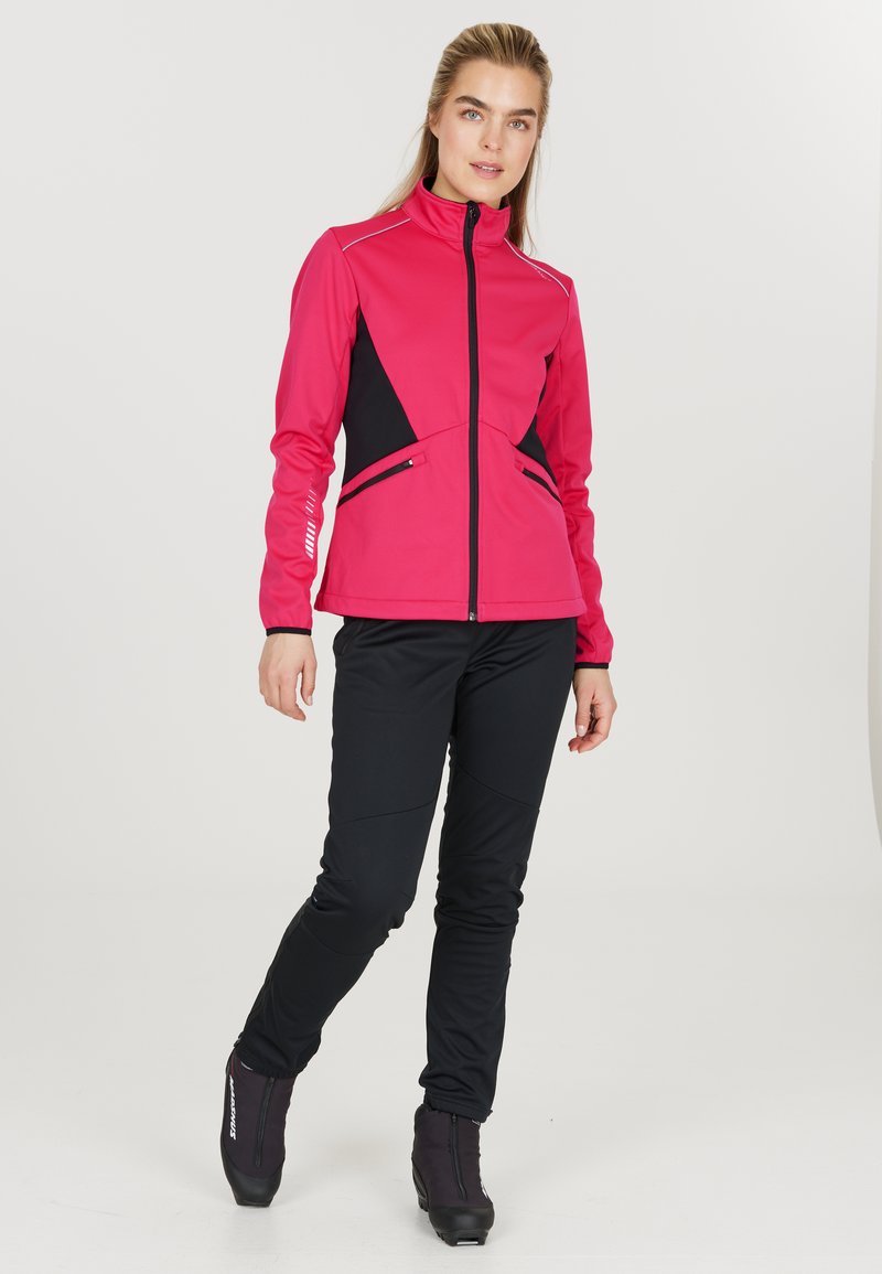 Fuchsia softshell-jakke med sorte accenter, høj krave, frontlynlås og sidelommer. Kombinationen med sorte tætsiddende bukser og sort fodtøj.