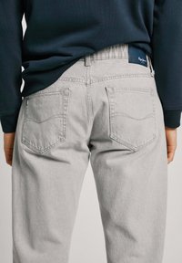 Pelēki denima džinsi ar atbrīvojošu piegriezumu, ar aizmugurējām kabatām, smalku šuvju detaļu un ar zīmola uzrakstu jostas daļā. Valkāti ar tumši zilu tops.