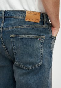 Sininen farkkukangas housut, joissa on ruskea nahkapaikka, jossa lukee "TOMMY JEANS", takataskuissa on erottuva ommel, ja pieni logotiedosto.