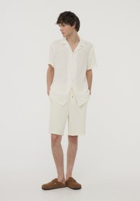 Camicetta beige chiaro a maniche corte realizzata in tessuto testurizzato, abbinata a pantaloni corti coordinati. Scarpe brown slip-on completano l'outfit. Il modello posa con calma.