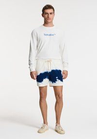 Camisa blanca de manga larga con el texto azul "Suncation™", acompañada de pantalones cortos color crema con un patrón de tie-dye azul y cintura con cordón. Zapatillas beige.