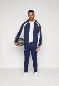 Jūras spēku sporta tērps ar baltajiem akcentiem, zip-up kapuco jaka un bikses ar elastīgu jostasvietu. Tur rokās melnu Spalding basketbola bumbu, kas rotāta ar izšūtām logo.