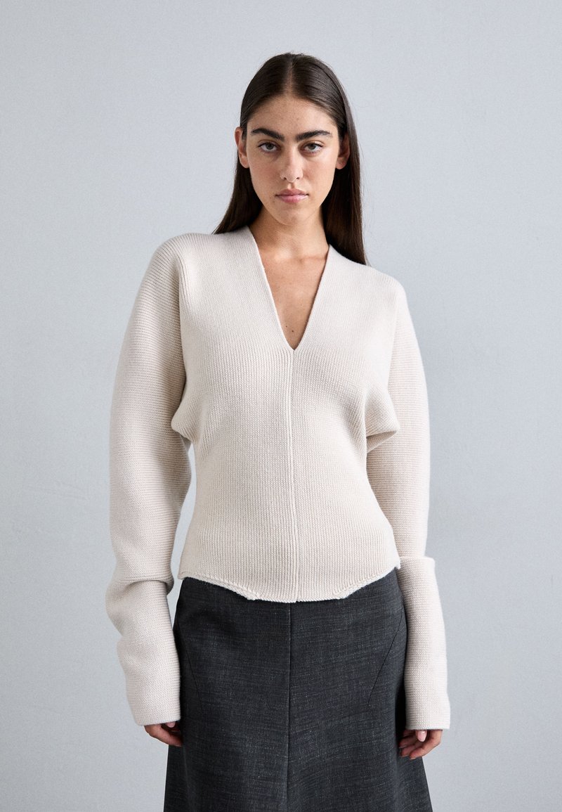 Bežs adīts džemperis ar v-veida kaklu, teksturēta auduma, ribotas struktūras un garām piedurknēm. Savienots ar tumši pelēku svārku.