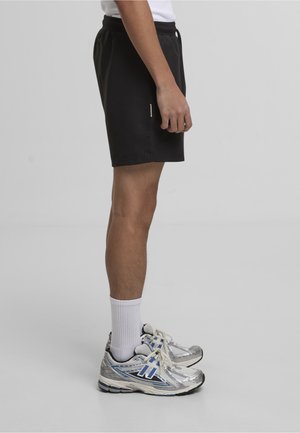 Hombre con pantalones cortos negros, calcetines blancos hasta la mitad de la pierna y zapatos deportivos plateados, de perfil sobre un fondo gris liso.