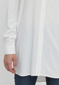 Weißes Langarmhemd mit Knopfleiste, weicher Textur, lockerer Passform und abgerundetem Saum, kombiniert mit dunkelblauen Jeans.