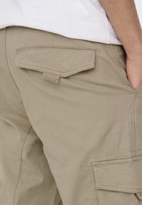 Kahe hlače od kaki pamučne tkanine s bočnim džepovima, pojačanim šavovima i teksturiranim završetkom. Sadrže stražnji džep s preklopom i detaljem gumba.