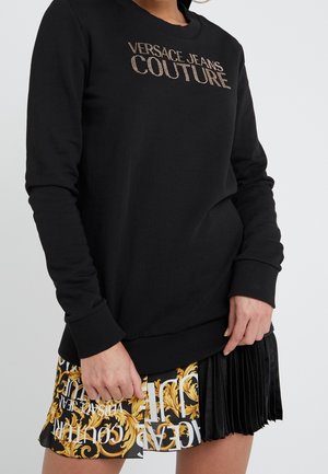 Czarna bluza z długim rękawem z "Versace Jeans Couture" z cyrkoniami, zestawiona z plisowaną spódnicą z motywem złotego baroku i białego tekstu.