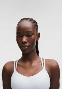 Top atletico alb cu bretele subțiri, ajustabile și decolteu în formă de cerc. Țesătură netedă și elastică cu un design strâns pe corp. Fundal neutru.