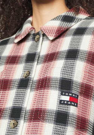 Gros plan d'une personne portant une chemise à carreaux rouge, noire et blanche avec des boutons marron et un patch logo Tommy Jeans sur la poche poitrine.