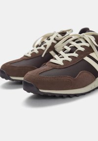 Brązowe sneakersy z zamszu i siateczki z kremowymi sznurówkami, z zaokrąglonym noskiem i teksturowaną podeszwą. Boki ozdobione kremowymi paskami.