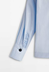 Poignet de chemise à manches longues bleu clair avec bouton décoratif noir et petit détail de volant en tissu près du poignet sur fond blanc.