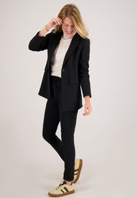 Zwarte blazer en bijpassende broek met een crèmekleurige top. Model draagt beige sneakers met zwarte strepen. Glad stof en een aangesloten pasvorm.